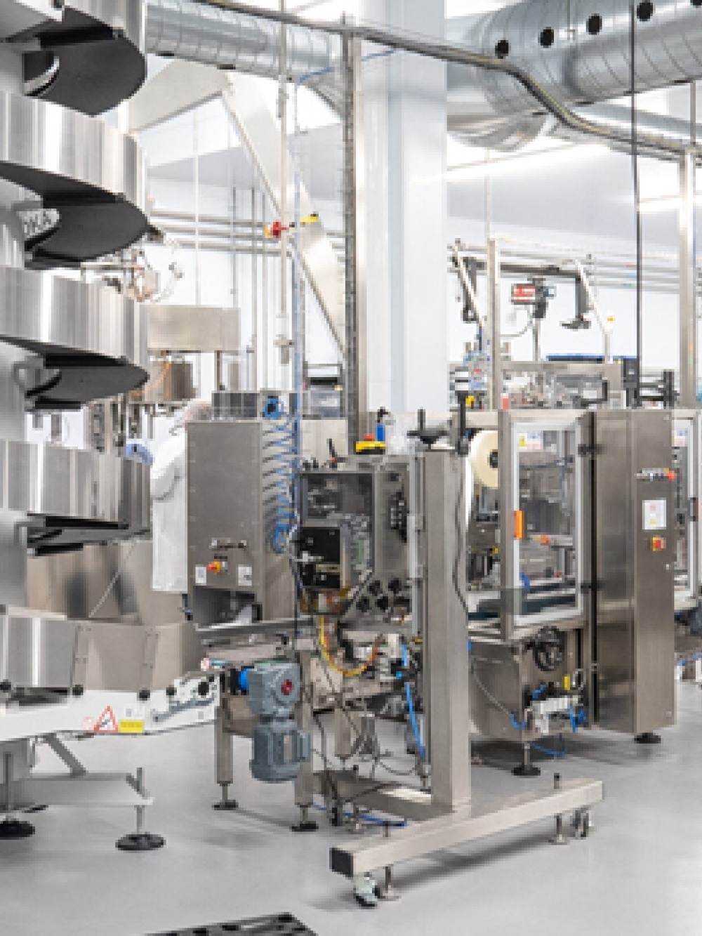 Salle de production industrielle avec des équipements automatisés pour l’embouteillage et le conditionnement, incluant des convoyeurs, des machines en acier inoxydable et des conduits aériens, dans un environnement propre et lumineux.