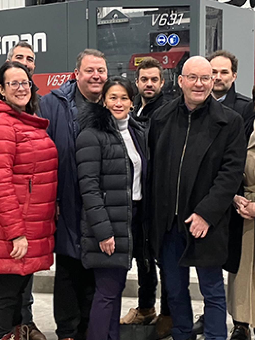 Groupe de personnes debout dans un atelier industriel devant une grande machine de production, portant des manteaux d’hiver et des vêtements de travail.