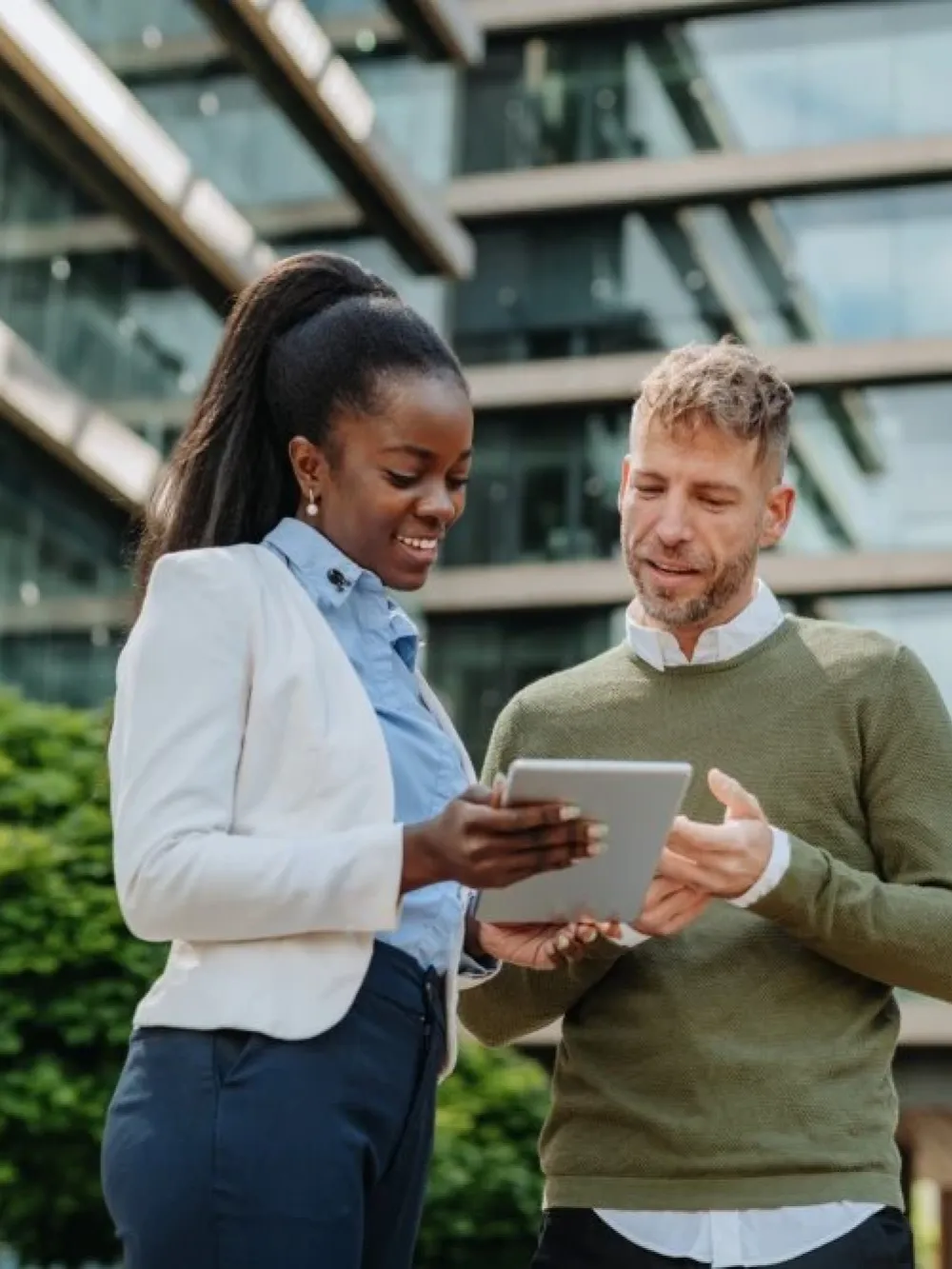 Deux personnes discutent ensemble à l’extérieur d’un immeuble moderne en consultant une tablette.