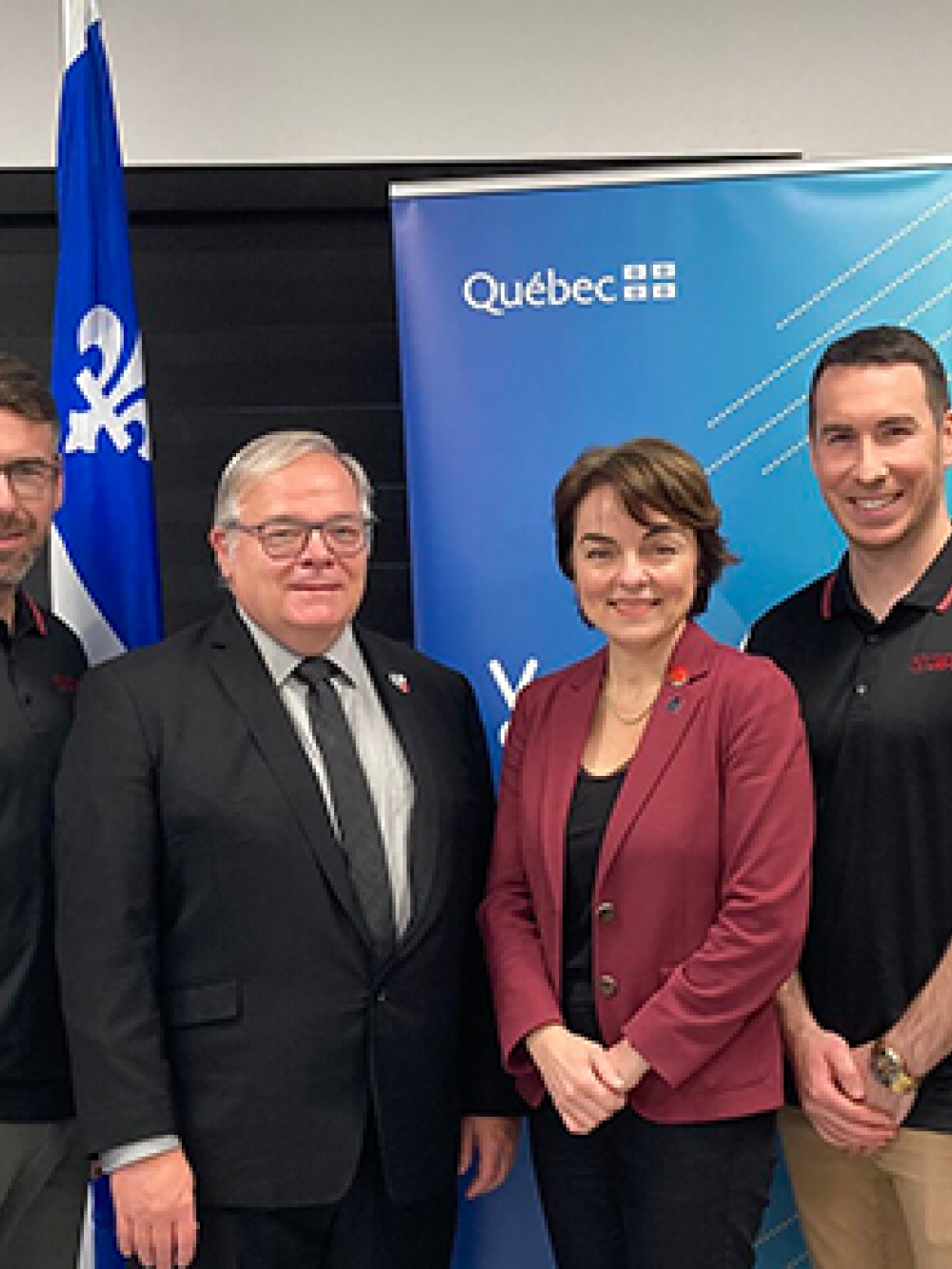 Groupe de personnes posant devant des bannières du gouvernement du Québec, incluant un drapeau fleurdelisé, dans une salle de présentation.
