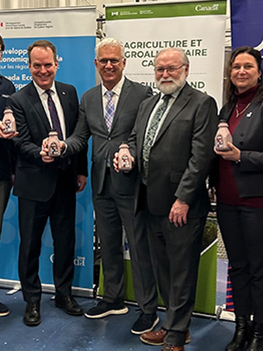 Groupe de sept personnes posant dans une usine devant des bannières gouvernementales, chacune tenant une petite bouteille blanche, avec des drapeaux du Québec et du Canada de chaque côté.