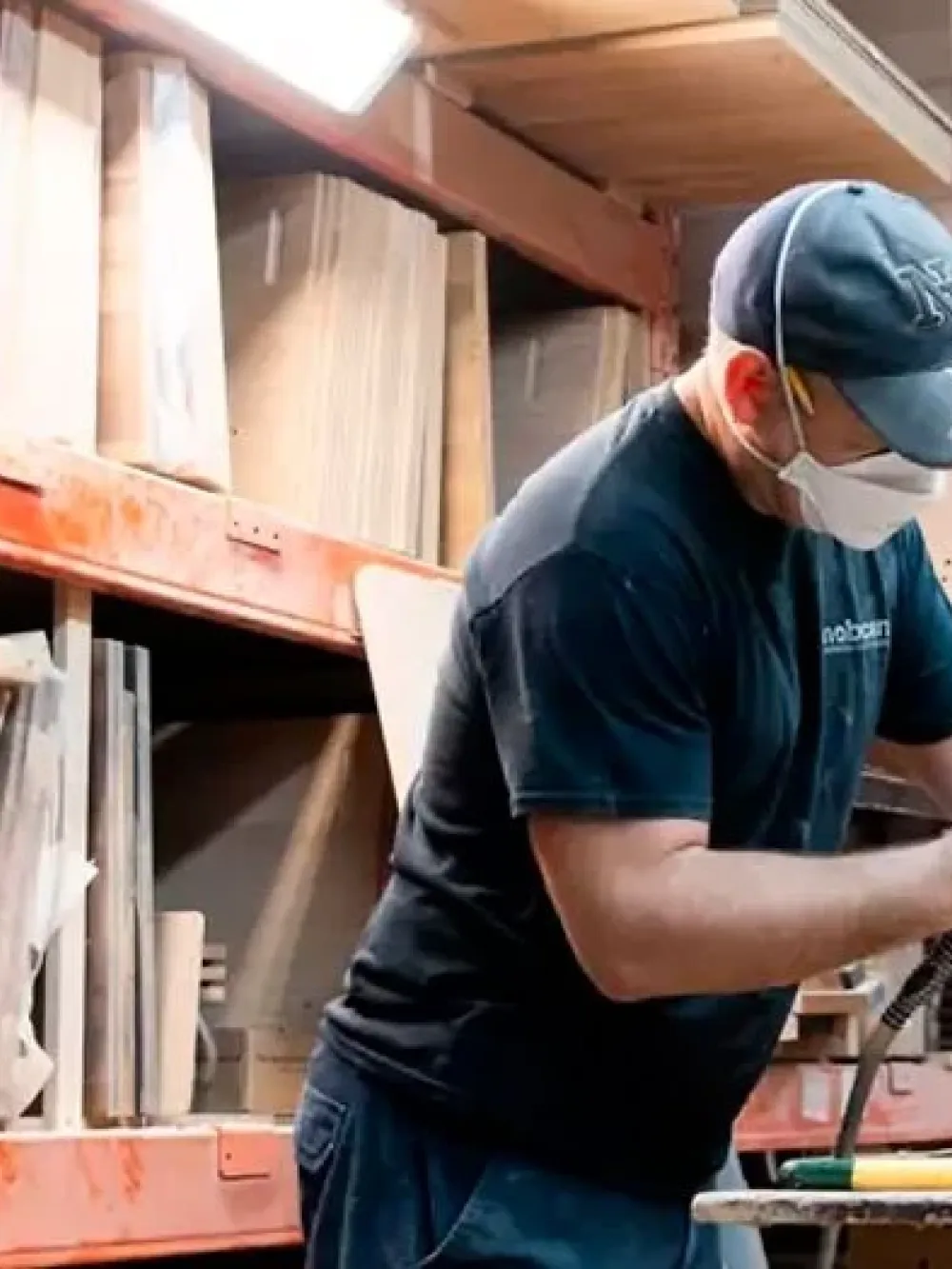 Un travailleur dans un atelier industriel, en train de poncer une pièce de bois sur un établi, entouré d’étagères contenant des panneaux et matériaux empilés.