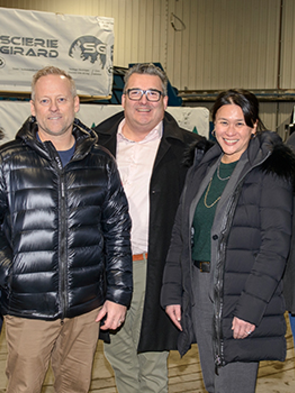 Groupe de personnes debout dans un entrepôt industriel, vêtues de manteaux d’hiver, posant devant des structures métalliques et du matériel de scierie.