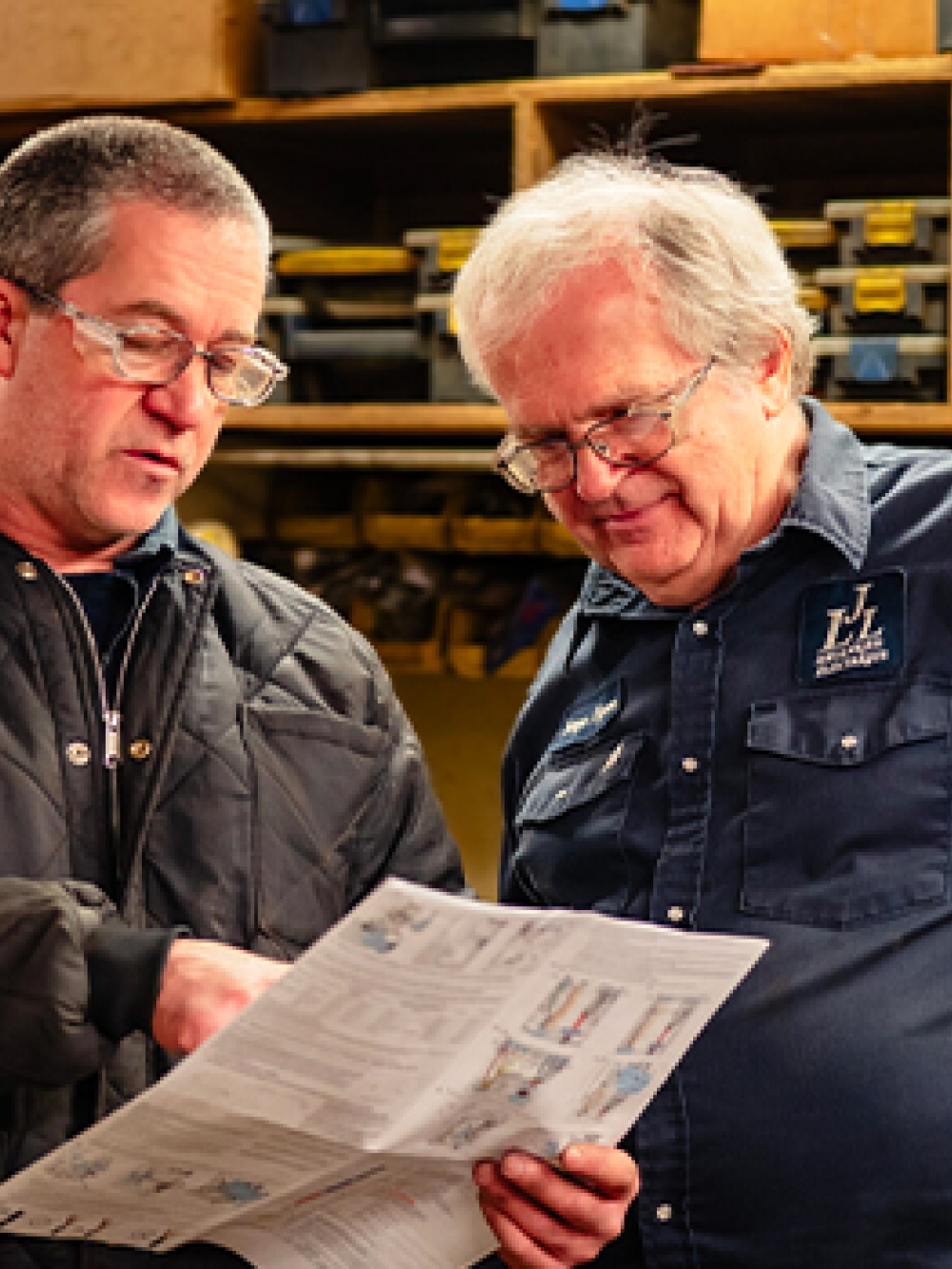 Deux personnes dans un atelier mécanique discutant et examinant un document technique, entourées d’étagères remplies de boîtes et d’outils.