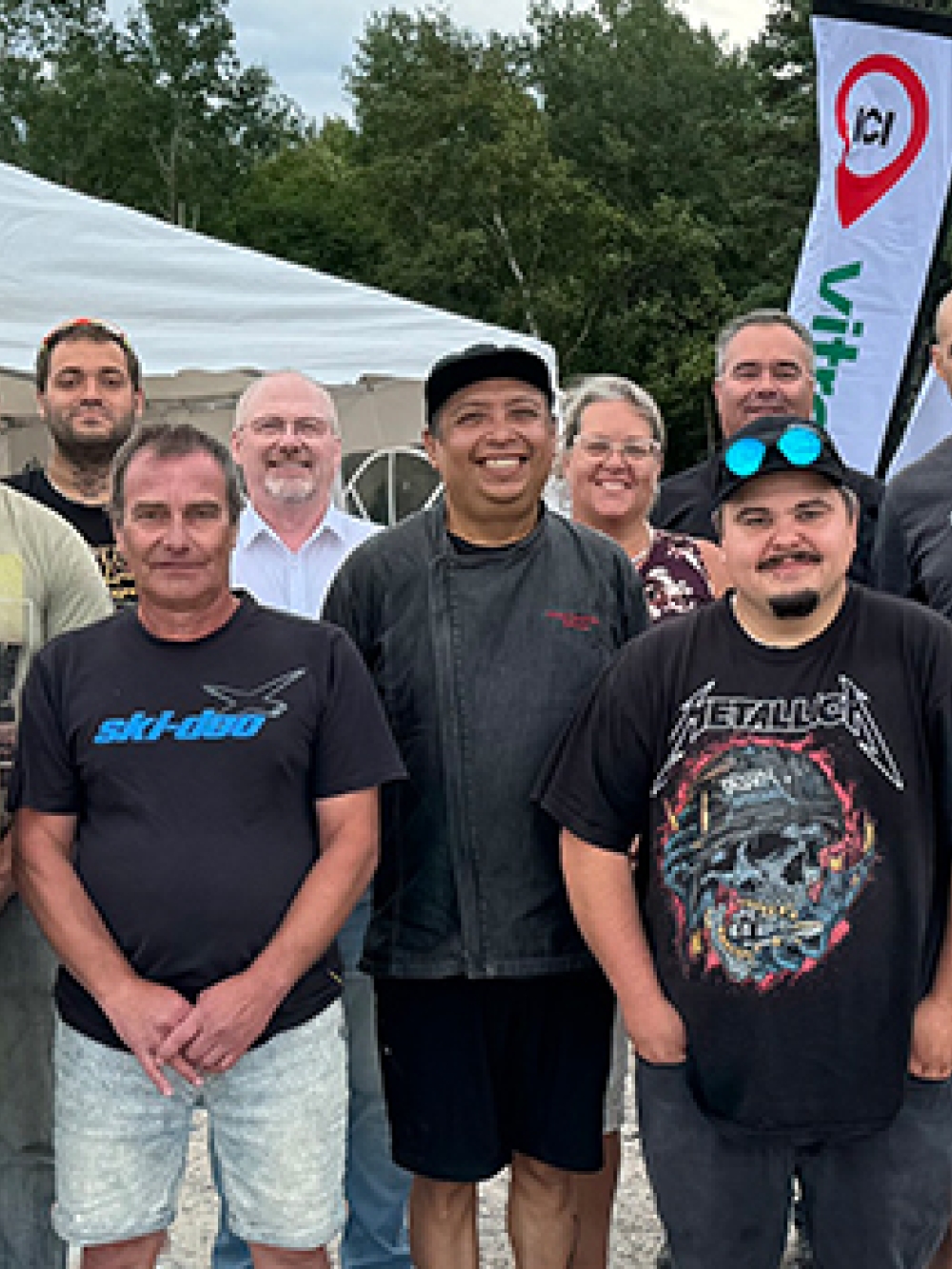 Groupe de personnes réunies à l’extérieur devant un chapiteau blanc, avec des bannières et une zone boisée en arrière-plan.