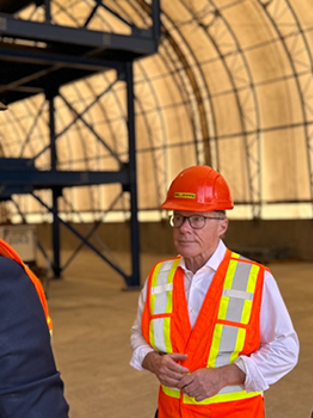 Trois personnes portant des casques et des vestes de sécurité orange discutent à l’intérieur d’un grand bâtiment industriel, près de structures métalliques et d’équipements.