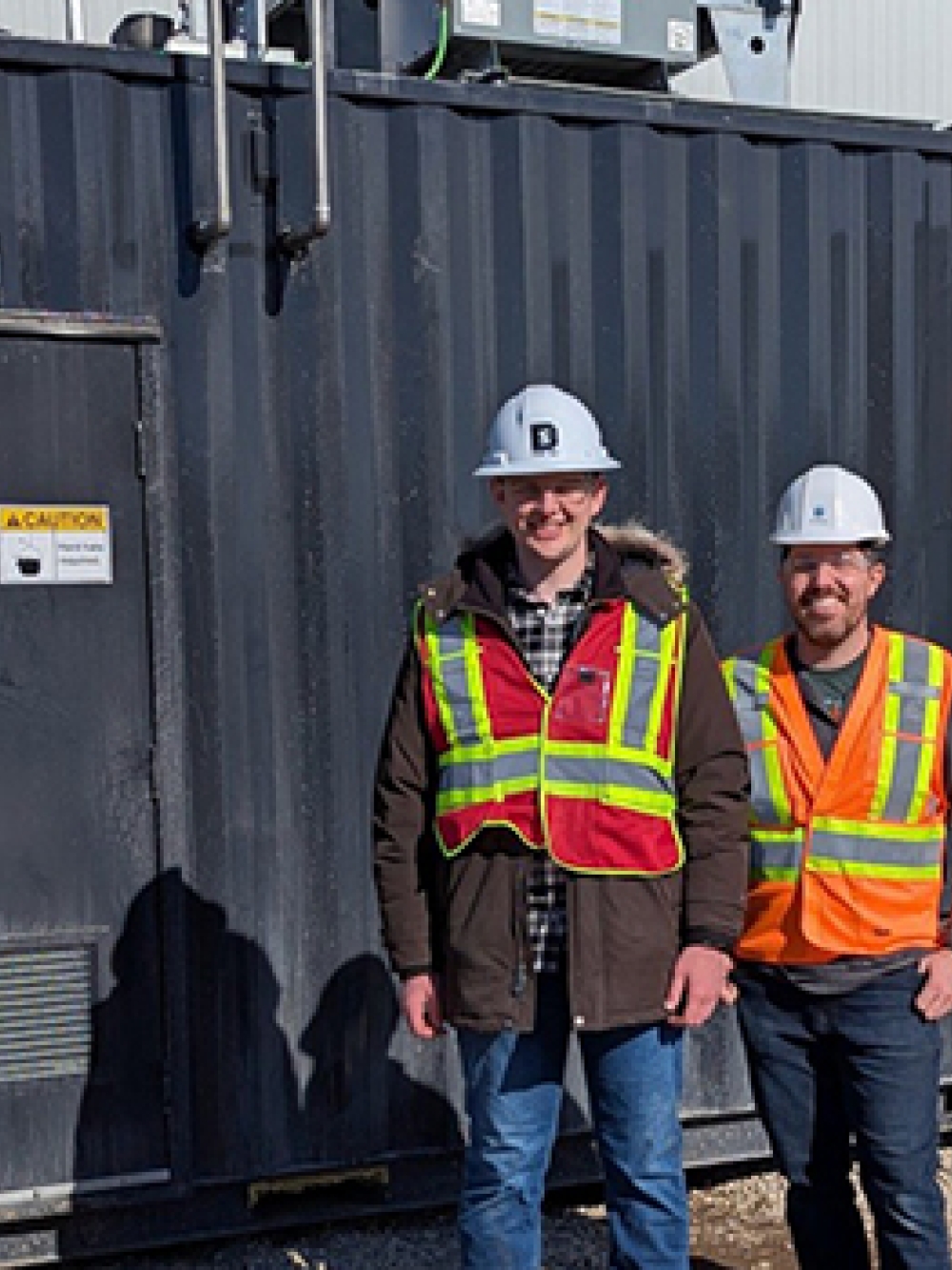 Trois personnes portant des casques de sécurité et des vestes haute visibilité debout devant une unité industrielle noire portant l’inscription « SKYRENU 50 » sur un site extérieur.