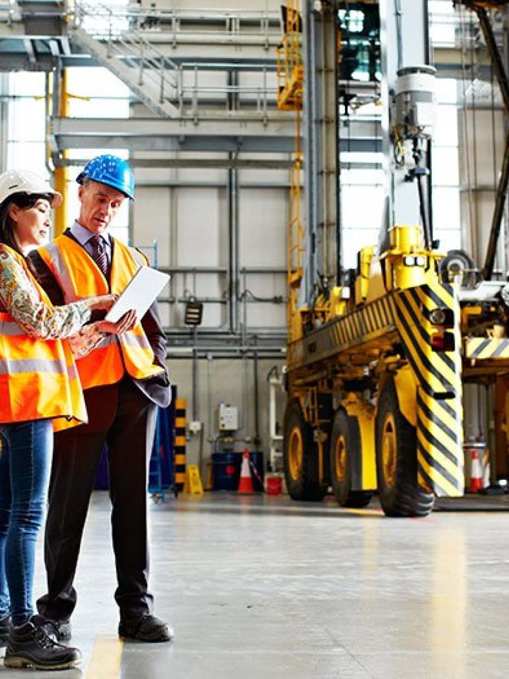 Deux personnes en gilet de sécurité et casque discutent dans un grand atelier industriel, debout devant de la machinerie lourde jaune et consultent des documents.