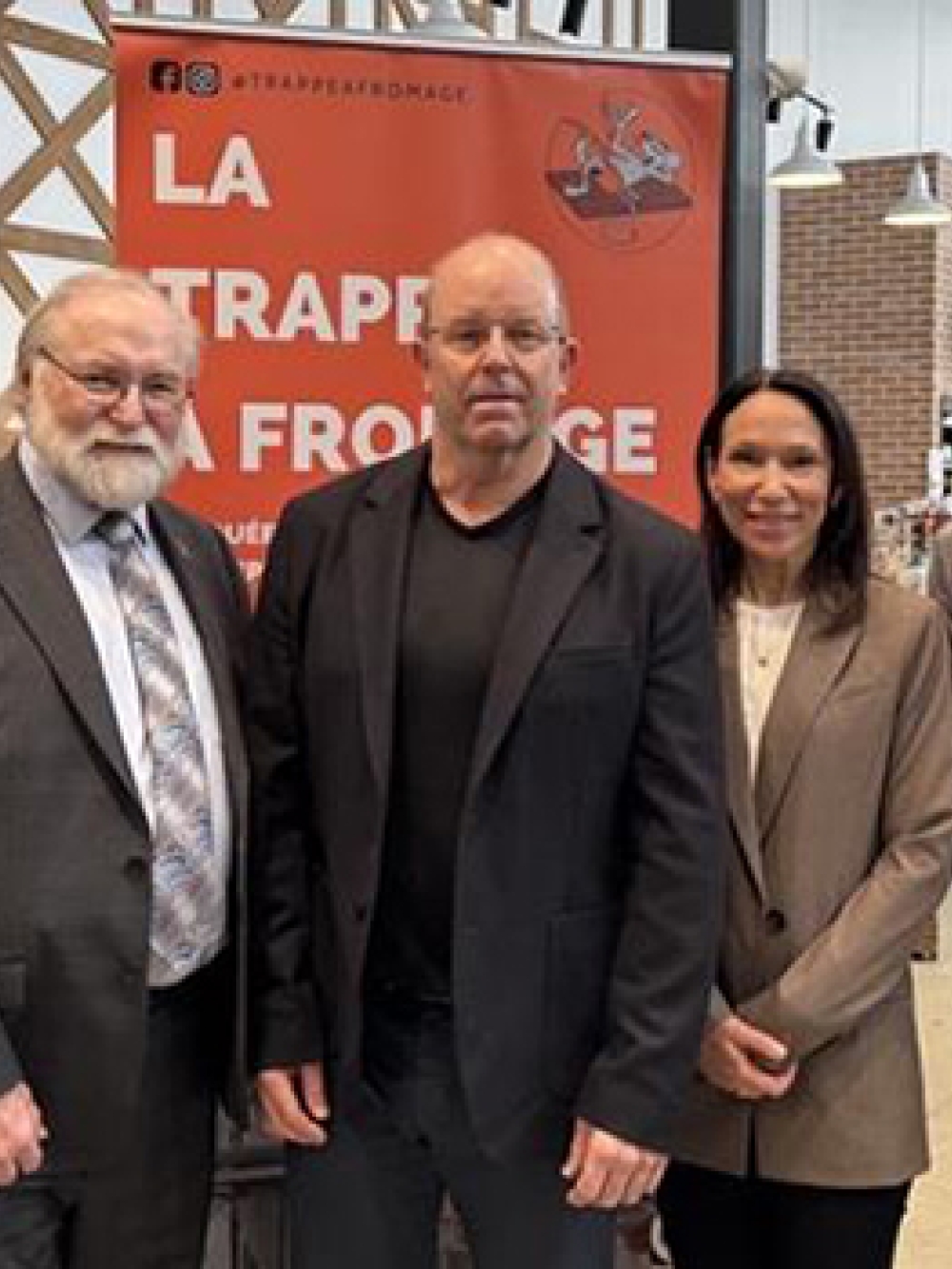 Groupe de cinq personnes posant dans un commerce de fromagerie, devant une affiche « La Trappe à Fromage » et un drapeau du Québec, avec des étagères de produits à droite.