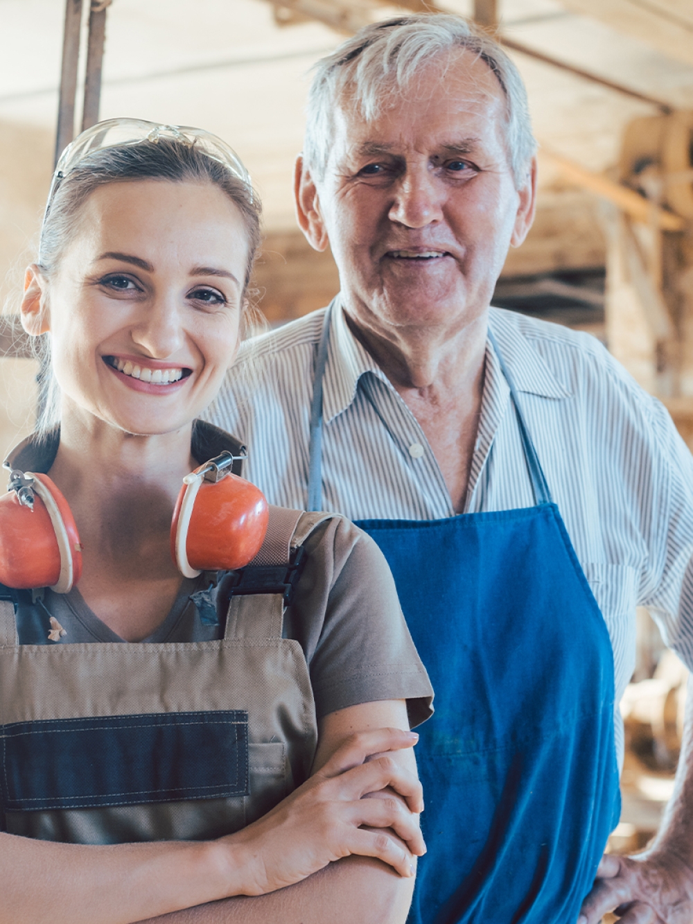 Jeune femme en tablier au premier plan d’un atelier de menuiserie, avec un homme en tablier à l’arrière‑plan, illustrant la continuité d’un savoir‑faire.